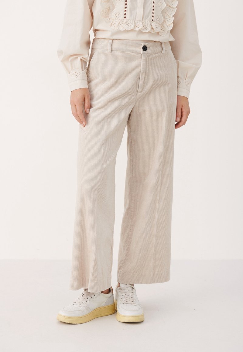 Pantalon large en velours côtelé beige porté avec des baskets blanches et un chemisier crème avec des détails en dentelle et œillets.