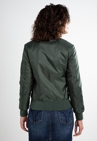 Oliwkowa kurtka bombowca z połyskującego nylonu, z prążkowanymi mankietami i dołem, posiadająca zamek błyskawiczny z przodu oraz kieszenie boczne. Pokazana z tyłu.