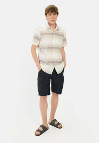 Kurzärmliges kariertes Hemd in Beige mit roten und blauen Streifen; kombiniert mit dunkelblauen Cargo-Shorts und schwarzen Sandalen. Lässiges Design und Passform.