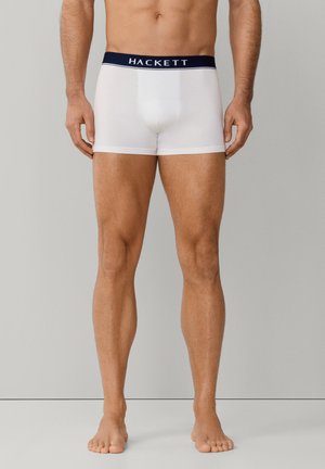 Mannelijke torso en benen in witte Hackett boxershorts met een marineblauwe tailleband, staand op blote voeten tegen een effen lichtgrijze achtergrond.