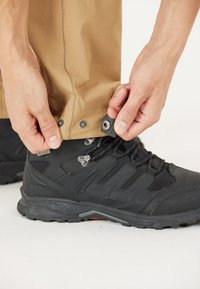 Zwarte waterdichte wandelschoen met een gestructureerd oppervlak en grijze accenten, naast beige broek die wordt dichtgeknoopt met drukknoopjes.