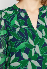 Bloemenblouse met een marineblauwe achtergrond en groene en crèmekleurige bladeren en bloemen, v-hals ontwerp en een zacht geplooide kraag.