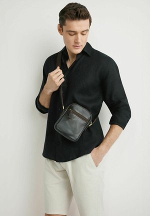 Mężczyzna w czarnej koszuli na guziki i jasnobeżowych szortach, niosący małą czarną skórzaną torbę na ramię, pozujący na tle białej ściany.