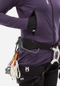 Personne portant une veste violette et un harnais d'escalade noir avec une corde et des mousquetons attachés à la ceinture du harnais.