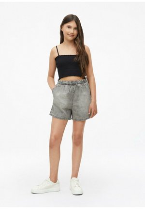 Junges Mädchen steht mit den Händen in den Taschen, trägt ein schwarzes Crop-Top, graue Jeansshorts und weiße Sneakers vor weißem Hintergrund.