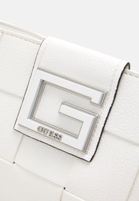 Hvid struktureret håndtaske med en sølvmetal logo-lås, der har et markant "G" og "GUESS" branding. Elegant design med syede detaljer.