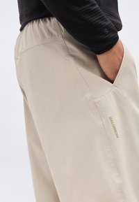 Άτομο που φοράει μπεζ παντελόνι Salomon με το χέρι στην πλαϊνή τσέπη, εμφανείς ραφές και μέρος μαύρης μακρυμάνικης μπλούζας.