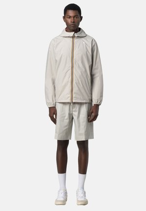 Uomo che indossa una giacca leggera beige con cappuccio e zip, pantaloncini coordinati fino al ginocchio, calzini bianchi e scarpe da ginnastica bianche, in piedi davanti a uno sfondo semplice.