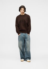 Νεαρός με σκούρα καστανά μαλλιά μέχρι τους ώμους, φοράει σκούρο καφέ φούτερ, φαρδιά μπλε τζιν και λευκά αθλητικά παπούτσια, στέκεται με τα χέρια στις τσέπες.