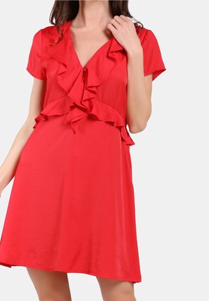 Frau trägt ein kurzes rotes Kleid mit gerafftem V-Ausschnitt und kurzen Ärmeln, posiert vor einem einfarbigen grauen Hintergrund.