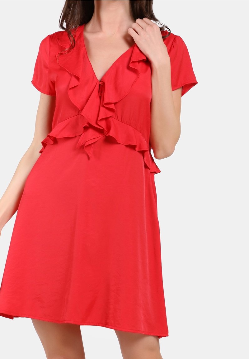 Femme portant une robe courte rouge avec un col en V à volants et des manches courtes, posant devant un fond gris uni.