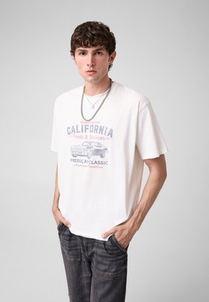 Giovane uomo con capelli ricci che indossa una t-shirt bianca con grafica e jeans scuri, in piedi con le mani in tasca davanti a uno sfondo semplice.
