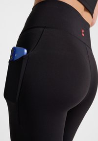 Svarta atletiska leggings med hög midja, med en sidoficka som rymmer en blå smartphone. Slät tyg med subtil textur.