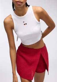 Top corto bianco con grafica di ciliegie, abbinato a una gonna rossa avvolgente. La gonna presenta un tessuto testurizzato e un orlo asimmetrico.