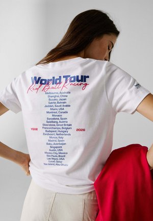 Naine, kes kannab valget Red Bull Racing World Tour 2026 t-särki, millel on loetletud ülemaailmsed võidusõidu asukohad, hoides punast jopet ühevärvilise tausta ees.