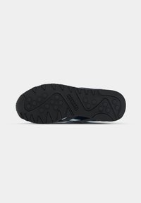 Semelle en caoutchouc noir de sneaker avec des motifs de grip texturés, featuring des rainures verticales et des points ronds. Le logo est embossé sur le côté.