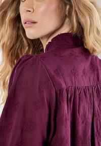 La blusa burdeos presenta un escote con volantes y patrones florales bordados en toda su superficie. La tela parece suave con un diseño de espalda fruncida.