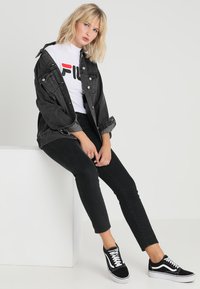 Veste en denim noir sur un t-shirt blanc avec un logo rouge et noir, associée à un jean skinny noir et des baskets noires à semelles blanches.