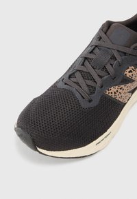 Baskets de course en maille noire avec un détail latéral à motif marron, lacets sombres et semelle blanche portant l'inscription "Fresh Foam" sur un fond blanc.