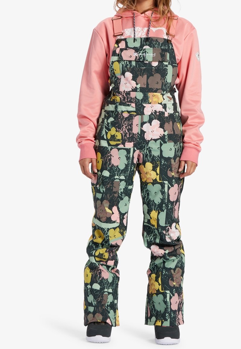 Person trägt Latzhose mit Blumenmuster in Grün-, Pink-, Gelb- und Brauntönen über einem pinkfarbenen Hoodie mit schwarzen und weißen Schuhen.