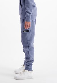 Persoon in blauwe cargobroek met zijzakken en witte sneakers, staande tegen een witte achtergrond.