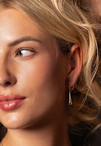 Boucles d'oreilles pendantes en ton or, avec une forme en goutte d'eau allongée et épurée, présentant une surface lisse et un design minimaliste.