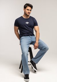 Granatowy bawełniany t-shirt z białym logo w kształcie diamentu na piersi, zestawiony z jasnoniebieskimi jeansami i czarnymi sneakersami.