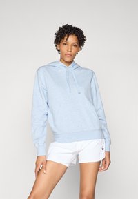 Felpa leggera blu con tasca frontale, caratterizzata da un cappuccio con cordino e logo ricamato. Abbinata a pantaloni sportivi bianchi.