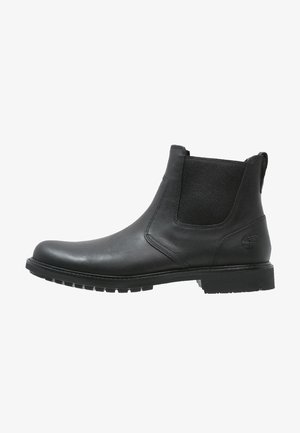 Bota Chelsea de cuero negro con panel elástico lateral, suela de goma y logo en relieve cerca del talón, vista desde el lado exterior.