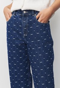 Jeansy dżinsowe w ciemnoniebieskim kolorze z subtelnym, białym wzorem w kokardki na całej powierzchni, z pięcioma kieszeniami oraz zapięciem na guziki.