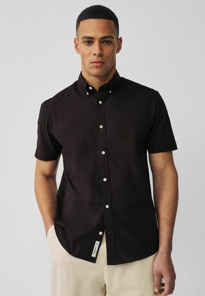 Egy férfi, aki egy testhezálló fekete, rövid ujjú, gombos inget visel krémszínű nadrággal, egy sima, világosszürke háttér előtt áll.