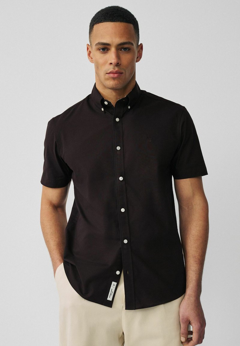 Homme portant une chemise noire cintrée à manches courtes avec des boutons, et un pantalon crème, debout devant un fond uni gris clair.