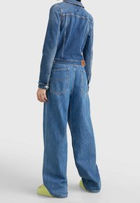 Person wearing blue denim jacket and wide-leg jeans, standing with back to camera, lime green platform sandals visible.