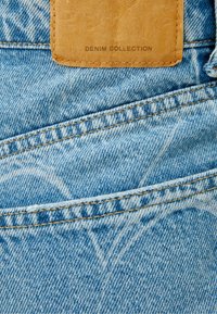 Ljust blå denimtyg med synliga sömmar, en brun läderlapp märkt "DENIM COLLECTION" och mjuka blekningsmönster.