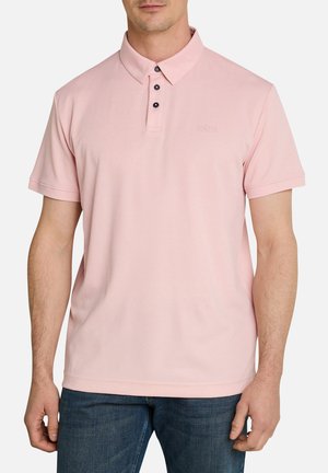 Homme portant un polo rose clair à manches courtes avec trois boutons noirs et un jean bleu, debout devant un fond blanc.