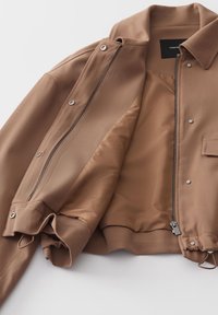 Bruine jas met een gladde buitenkant en een zachte voering. Voorzien van een ritssluiting aan de voorkant, drukknoopsluitingen en een trekkoord aan de onderkant. Geen patronen.