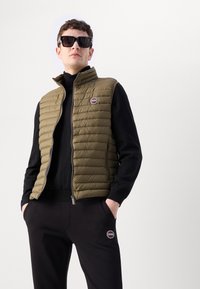 Colete acolchoado verde oliva com zíper, gola alta, bolsos laterais e um emblema circular. Usado sobre um suéter preto e calças pretas.