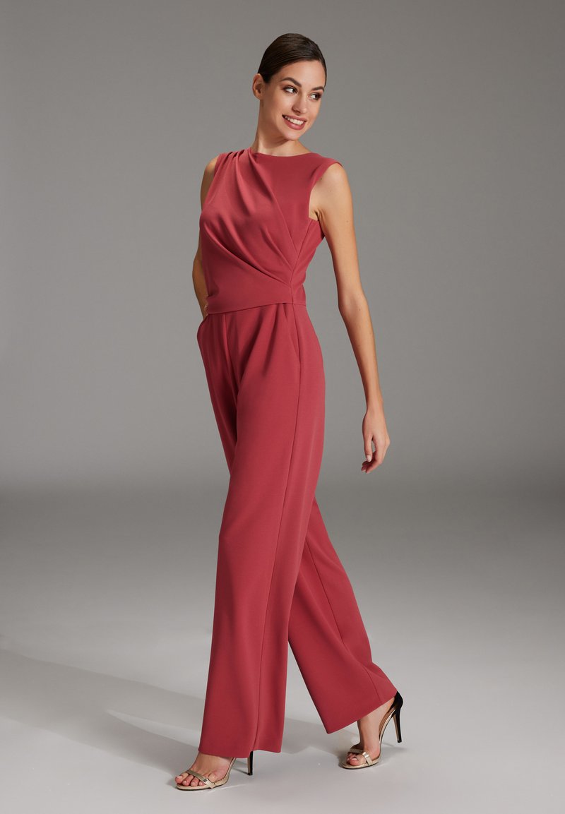 Szkarłatny kombinezon bez rękawów z draperią w talii, wysokim stanem i szerokimi nogawkami. Noszony z sandałami na obcasie.