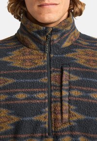 Pull semi-zippé en polaire bleu marine avec des motifs en diamants et des rayures en orange brûlé et jaune. Doté d'un col montant et d'une fermeture éclair.