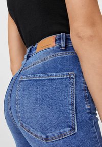 Jeans en denim bleu taille haute avec une finition texturée, présentant une pièce en cuir avec le nom de la marque, des poches arrière et des surpiqûres en contraste.
