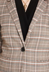 Blazer à carreaux marron avec un design structuré. Comprend un bouton noir et des revers prononcés, superposé sur un vêtement noir en dessous.