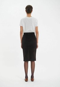 Jupe crayon en velours côtelé noir avec fente arrière, associée à un T-shirt gris clair. Des collants noirs transparents et des chaussures marron complètent le look.