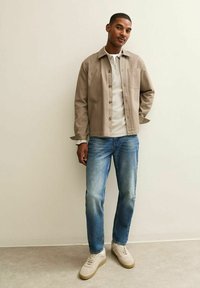 Chaqueta de botón marrón claro sobre una camisa blanca, combinada con jeans azul desgastados y zapatillas beige, de pie contra un fondo neutro.