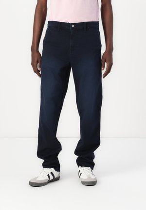 Mann trägt dunkelblaue Freizeithose, weiße Sneakers mit schwarzen Streifen und ein hellrosa Hemd, steht vor weißem Hintergrund.