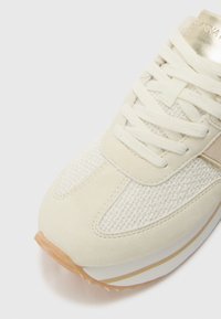 Chaussure de sport couleur crème comprenant un mélange de tissu texturé et de daim doux, avec une semelle en caoutchouc et de subtils accents dorés.