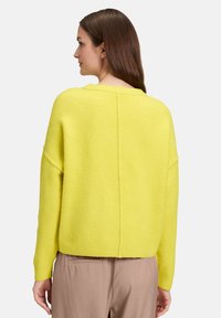 Kobieta z długimi brązowymi włosami w jasnosłonecznym żółtym swetrze i beżowych spodniach, pokazana od tyłu na tle gładkiego jasnego tła.