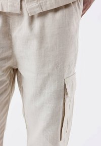 Beige linned cargo bukser med en tekstureret overflade, sidelommer og diskrete broderede detaljer på benet. Afslappet pasform.