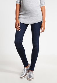 Mörkblå skinny jeans med en slät denimtextur, tillsammans med silverfärgade oxfordskor. Toppdelen är en ljusgrå, randig, långärmad tunika.