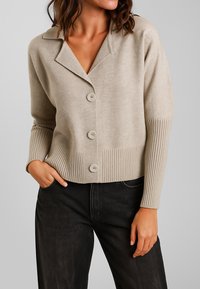 Donna in cardigan beige con bottoni e polsini e orlo a coste, abbinato a jeans neri, con una mano in tasca, su uno sfondo neutro.