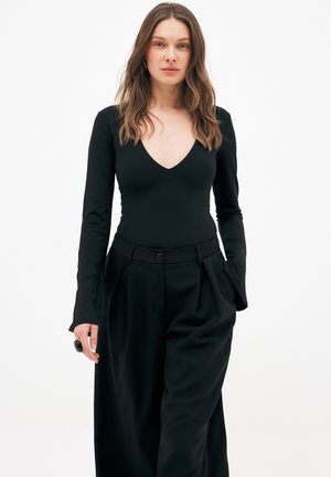 Žena s vlnitými hnedými vlasmi, oblečená v priliehavom čiernom top s véčkovým výstrihom a dlhými rukávmi, a vysokými čiernymi širokými nohavicami, ruky v vreckách.